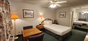 A hotel room with a bed, a desk, a chair, a nightstand, a lamp, a ceiling fan, and a bathroom with a sink and mirror. - Formosa Inn (Fresno)