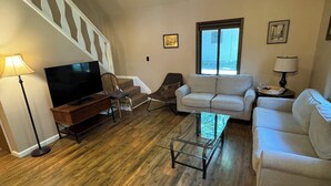 A spacious living room with a wooden floor, a flat-screen TV, a glass coffee table, and two white sofas.