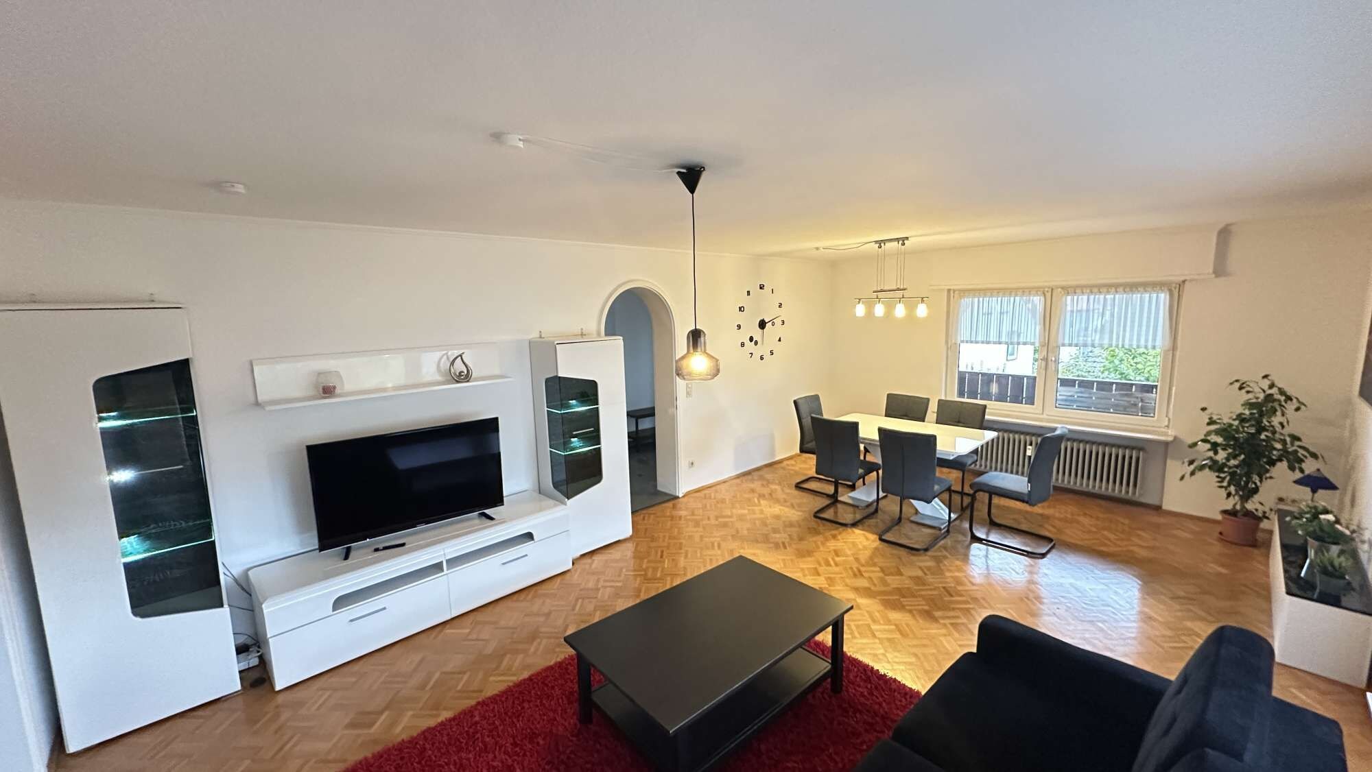 Ein modernes Wohnzimmer mit einem Flachbildfernseher, einem Essbereich mit Tisch und Stühlen sowie einem roten Teppich.