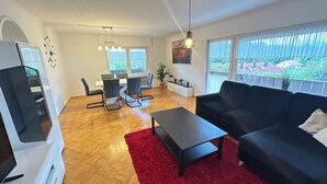 Een moderne woonkamer met een bank, salontafel en televisie. Er is een eethoek met stoelen en een tafel, en grote ramen met gordijnen.
