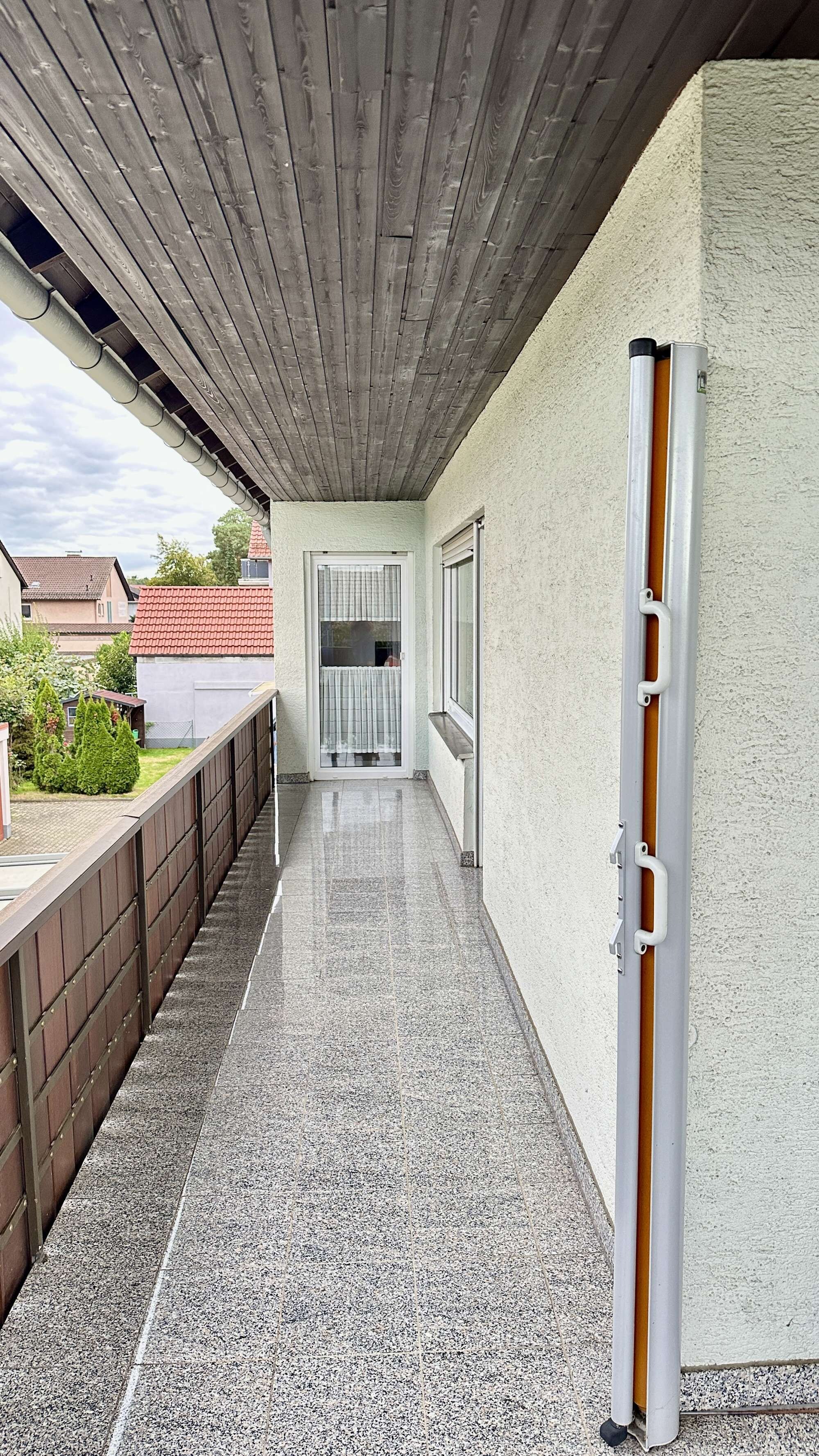 A covered outdoor corridor with a tiled floor, a wooden ceiling, and a view of a residential area in the background.