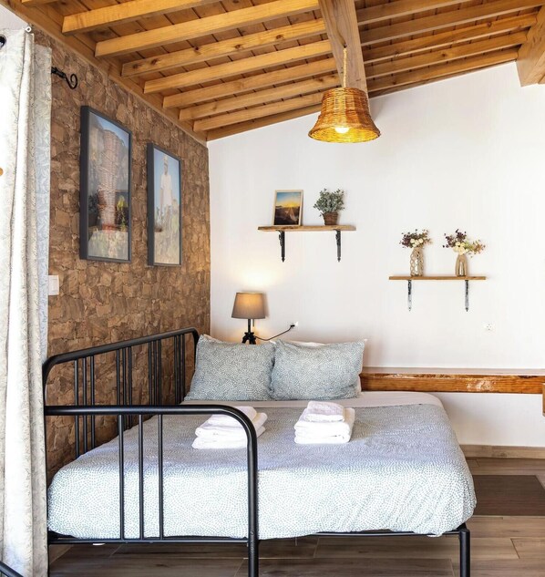 Ein Schlafzimmer mit einem Bett, einer Steinwand, Holzbalken, einer Pendelleuchte und Regalen an der Wand mit Pflanzen.