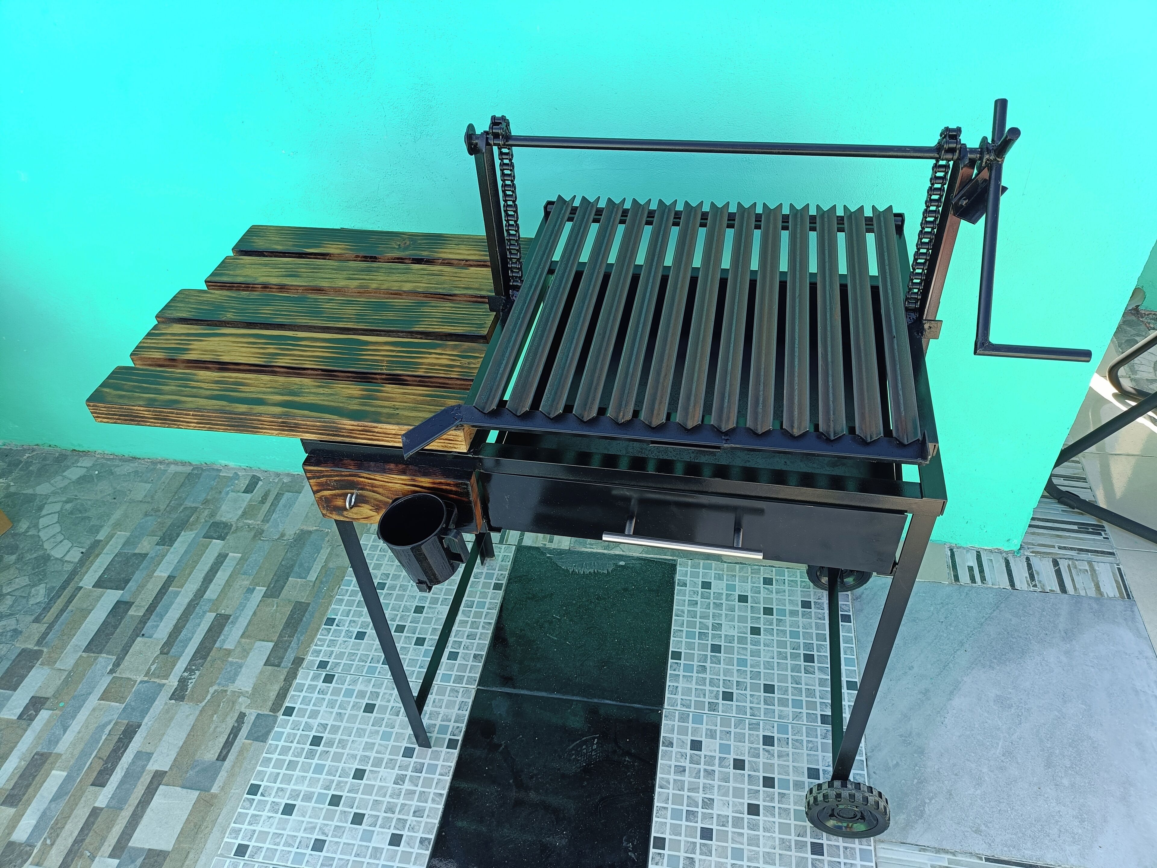 A metal grill with a wooden top and a metal base, placed on a tiled floor against a turquoise wall.