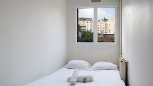 A small, white, single-bed room with a window overlooking a cityscape. - Appartement Lavoisier - Welkeys (Puteaux)