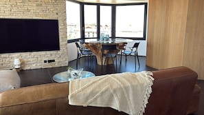 A modern living room with a flat-screen TV mounted on a brick wall, a round wooden table with chairs, and a large window offering a view of buildings.