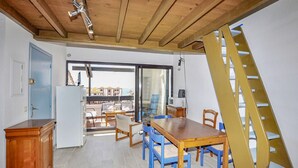 A room with a wooden ceiling, a dining table with blue chairs, a wooden cabinet, and a view through a glass door to an outdoor area with a chair and a table. - Amazing apartment in Le Barcarès (Le Barcarès)