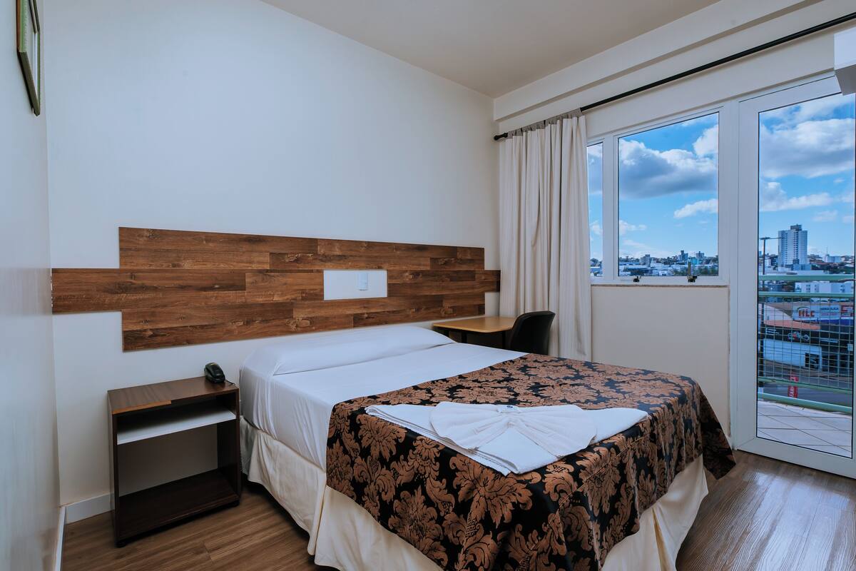 A hotel room with a wooden headboard, a large bed with a patterned bedspread, a bedside table, and a view of the city through a large window.