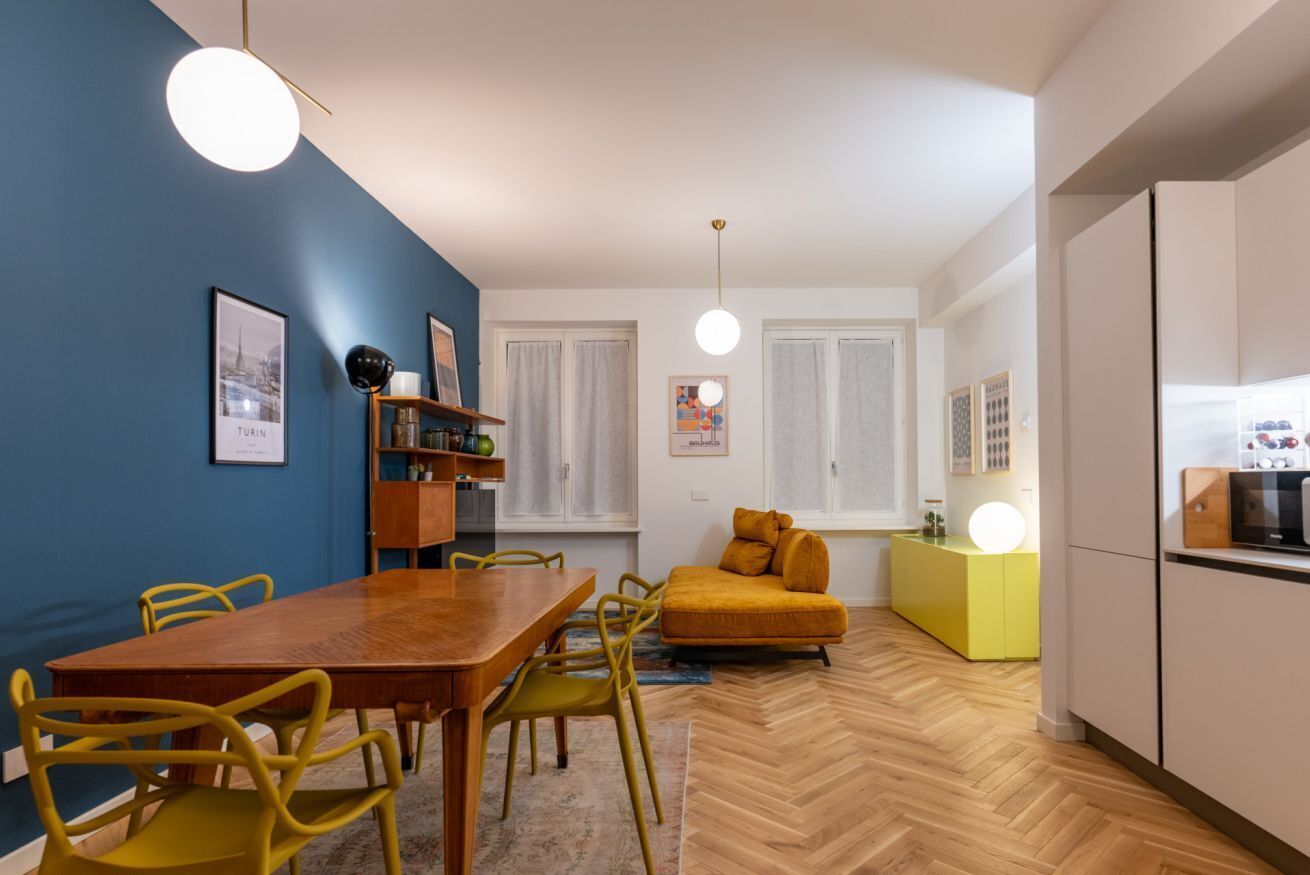 A dining area with a wooden table and yellow chairs, a sofa, a yellow cabinet, and a kitchen area with a refrigerator.
