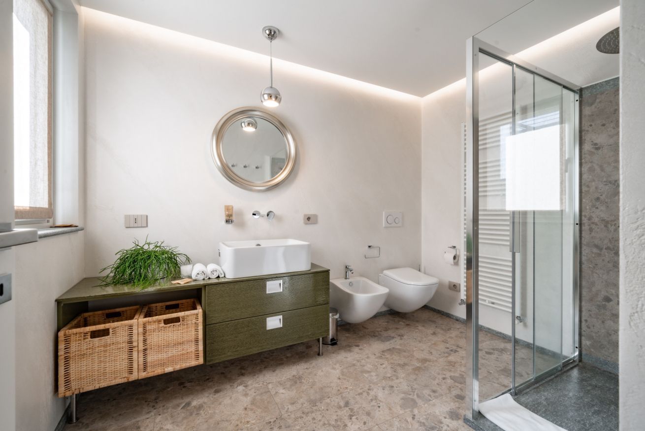 A modern bathroom with a glass shower enclosure, a round mirror, a green vanity with wicker baskets, and a bidet.