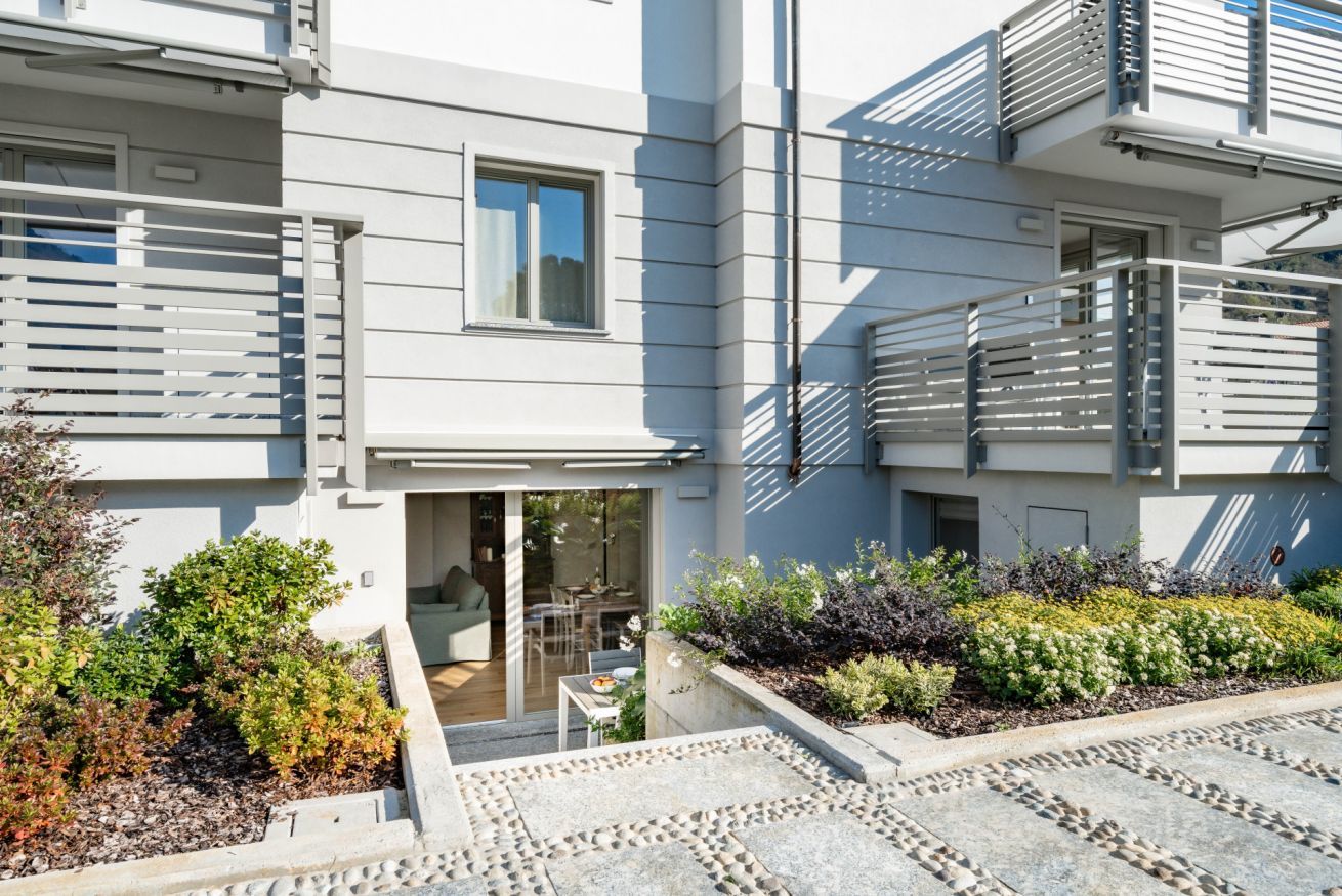 A modern apartment building with balconies, a patio area with seating, and a variety of plants.