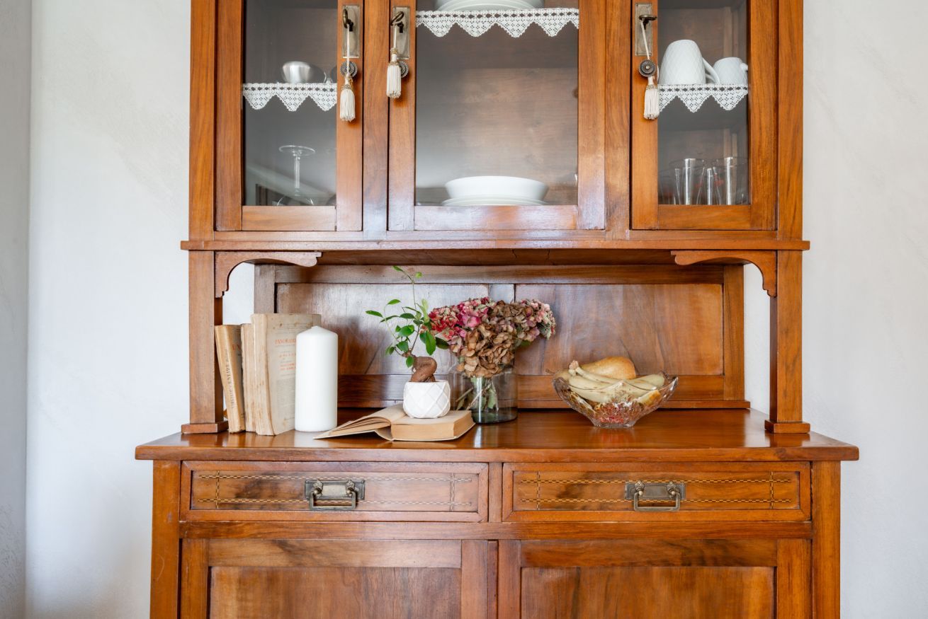 Un meuble en bois avec des portes en verre, un vase contenant des fleurs séchées et un bol de pain sur le comptoir.