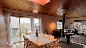 A dining area with a wooden table and chairs, a hanging light fixture, and a view of the outdoors through large windows.
