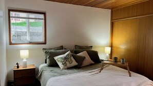 A bedroom with a bed, pillows, a bedside table with a lamp, a window with a view, and a wooden wardrobe.