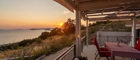 Terraço com mesa e cadeiras, com vista para a costa ao pôr do sol.