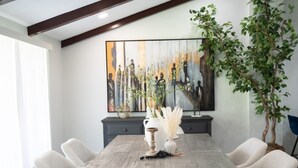 A dining room with a rustic wooden table, six white chairs, a large abstract painting on the wall, and a potted plant.