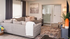 A living room with a grey sofa, a glass door leading to a bathroom, a ceiling fan, and a small table with a lamp.