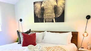 A hotel room with a large bed, a headboard, a wall-mounted elephant artwork, a bedside table with a lamp, and a neatly made bed with a patterned throw blanket.