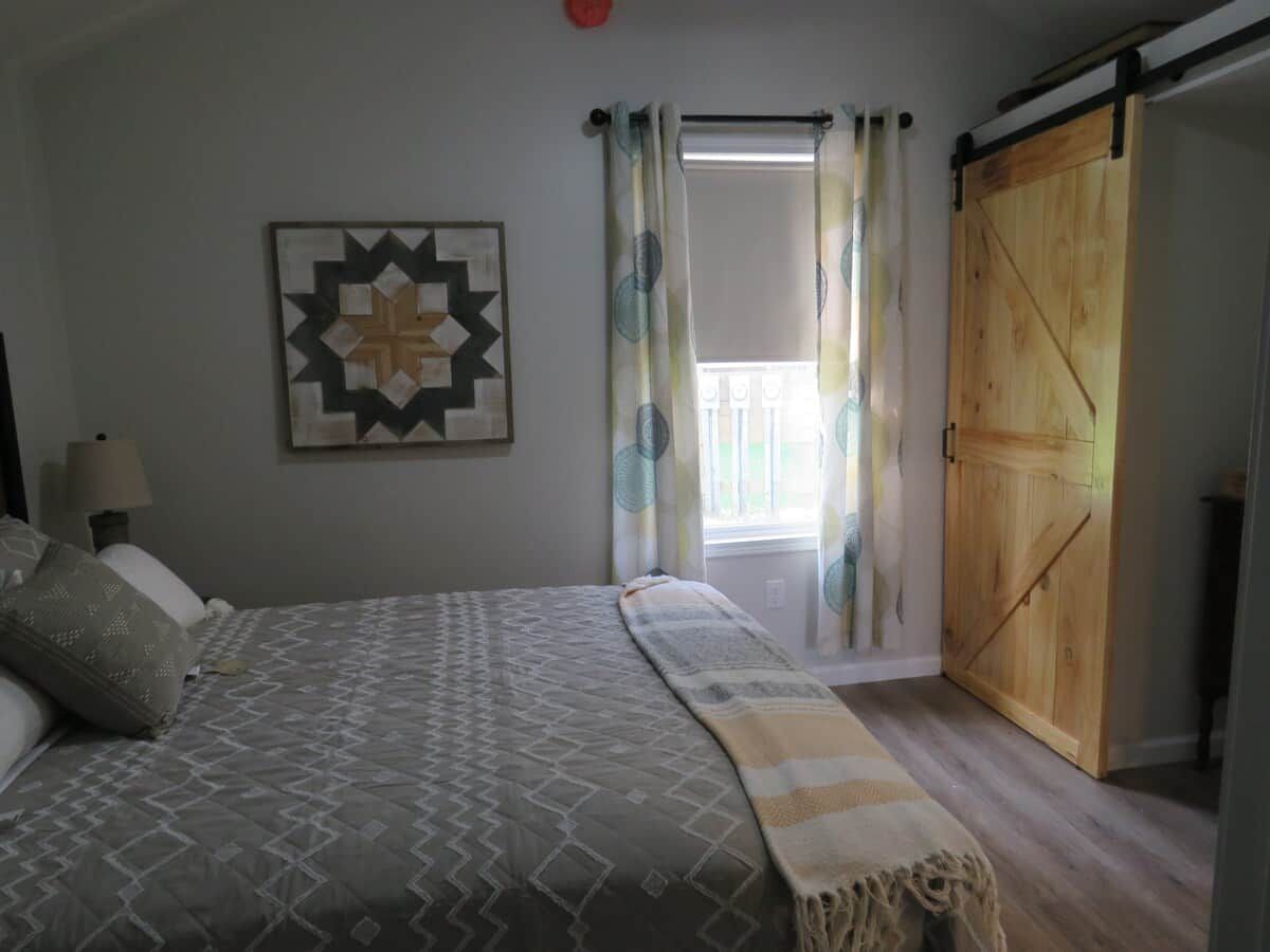Un dormitorio con cama, una obra de arte en la pared, una ventana con cortinas y una puerta de madera.