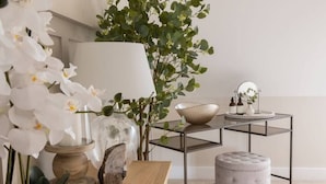 Ein modernes Interieur mit einem Holztisch, einer Glasvase mit weißen Blumen, einem Spiegel und einem kleinen Hocker.