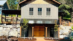 A two-story wooden cabin with a garage, surrounded by rocks and trees.