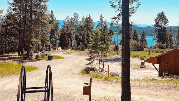 A rustic cabin with a dirt path leading to a wooden structure, surrounded by tall pine trees and a clear blue sky.