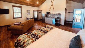 A spacious living room with a high ceiling, wooden beams, a large sofa, a patterned rug, and a kitchen area with modern appliances.