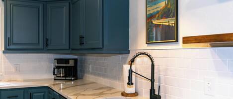 A kitchen with teal cabinets, a white tiled backsplash, a stainless steel sink, and a coffee maker.
