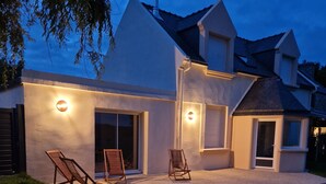 Une maison moderne dotée d’une terrasse en gravier, de deux fauteuils de jardin et d’un chemin menant à l’entrée.