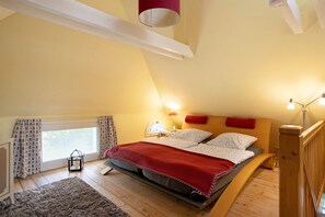 Une chambre avec un plafond en pente, un lit recouvert d’une couverture rouge, deux lampes de chevet et une fenêtre avec des rideaux.