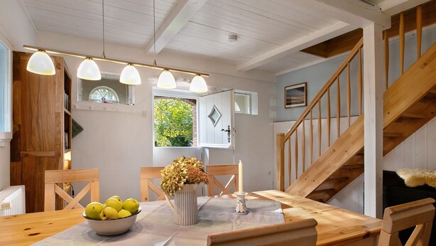 Un coin repas avec une table en bois, des chaises, un saladier de fruits, un vase de fleurs et un escalier.