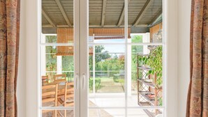 Ein Zimmer mit einer Schiebetür zu einem Außenbereich mit einem Patio-Tisch und -Stühlen, Blick auf einen Garten und einem Metall-Pflanzgefäß.