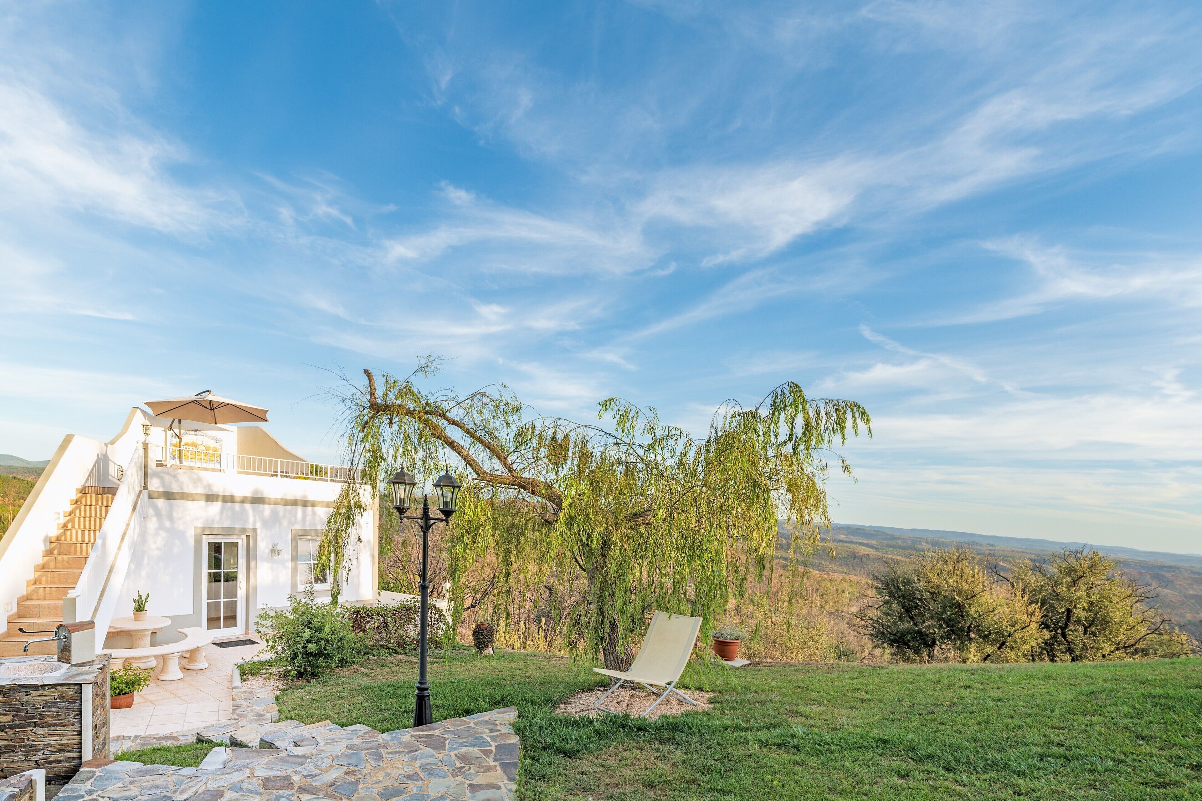 Una casa blanca con una escalera de piedra, un patio con mesa y sillas, una silla de jardín y un sauce llorón.