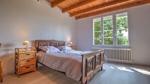 A bedroom with a wooden bed, two bedside tables with lamps, a radiator, and a large window with a view of trees. - Le Gîte du Sourcier – Authentic and Welcoming Stay in Dordogne (Sadillac)