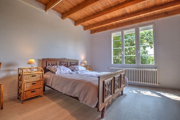 Un dormitorio con una cama de madera, dos mesitas de noche con lámparas, un radiador y un ventanal con vistas a árboles.