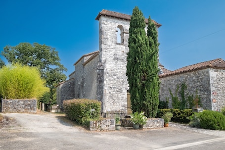 A stone building with a tower, surrounded by greenery and a gravel driveway.. Le Gîte du Sourcier – Authentic and Welcoming Stay in Dordogne