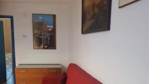Een rode bank in een kamer met een betegelde vloer, een houten kast en twee ingelijste foto's aan de muur.