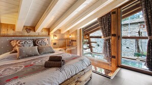 Ein Schlafzimmer in einer Holzhütte mit einem Bett, Kissen und einem Fenster mit Blick auf ein Steinhaus.