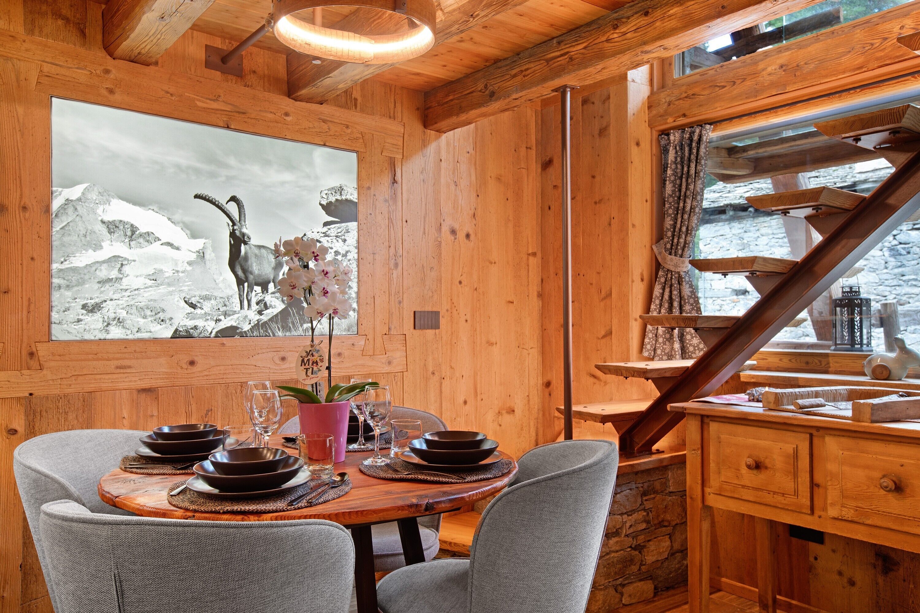 Un intérieur en bois avec une table à manger dressée pour quatre personnes, un tableau représentant un paysage de montagne et un escalier menant à un balcon.