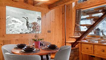 Un intérieur en bois avec une table à manger dressée pour quatre personnes, un tableau représentant un paysage de montagne et un escalier menant à un balcon.