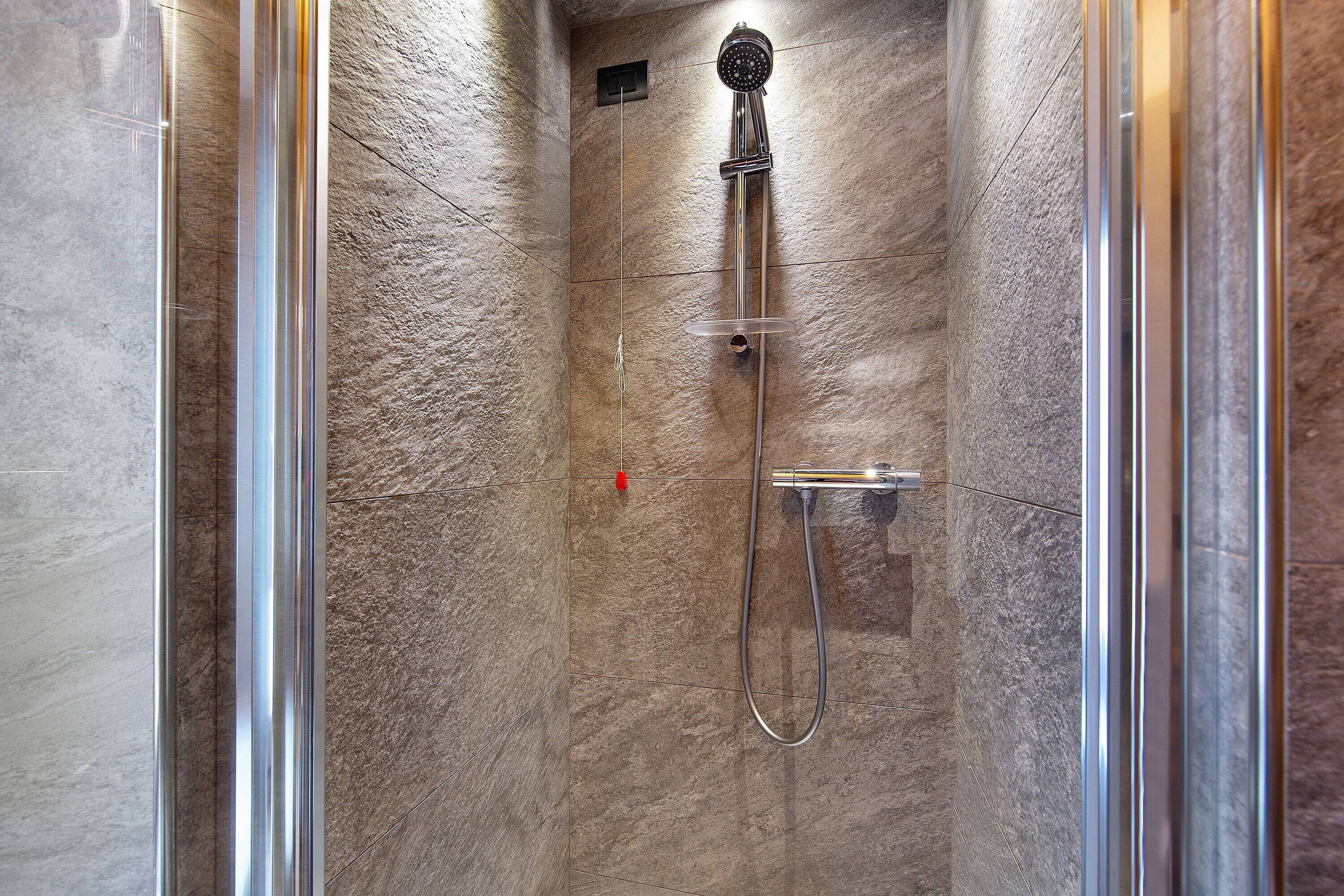 Une douche équipée d’un pommeau de douche à main et d’un robinet mural, installée dans une pièce aux murs en pierre et dotée d’une porte en verre.