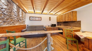 Un interior rústico con pared de piedra, vigas de madera en el techo, un sofá, una mesa de comedor y una zona de cocina.