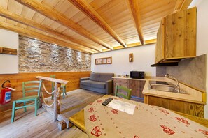 A rustic kitchen with wooden beams, a stone wall, a sofa, and a table with a remote control. - Apartment 'Veleza' with Mountain View and Shared Garden (Auronzo di Cadore)