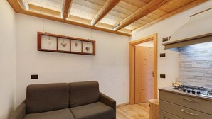 A room with a sofa, wooden ceiling beams, a framed picture, a wooden door, and a kitchen area with a stove. - Apartment 'Ciariè' with Mountain View (Auronzo di Cadore)