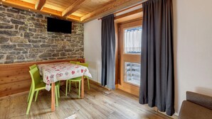 A room with a stone wall, wooden ceiling beams, a dining table with chairs, a television mounted on the wall, and a window with curtains. - Apartment 'Ciariè' with Mountain View (Auronzo di Cadore)