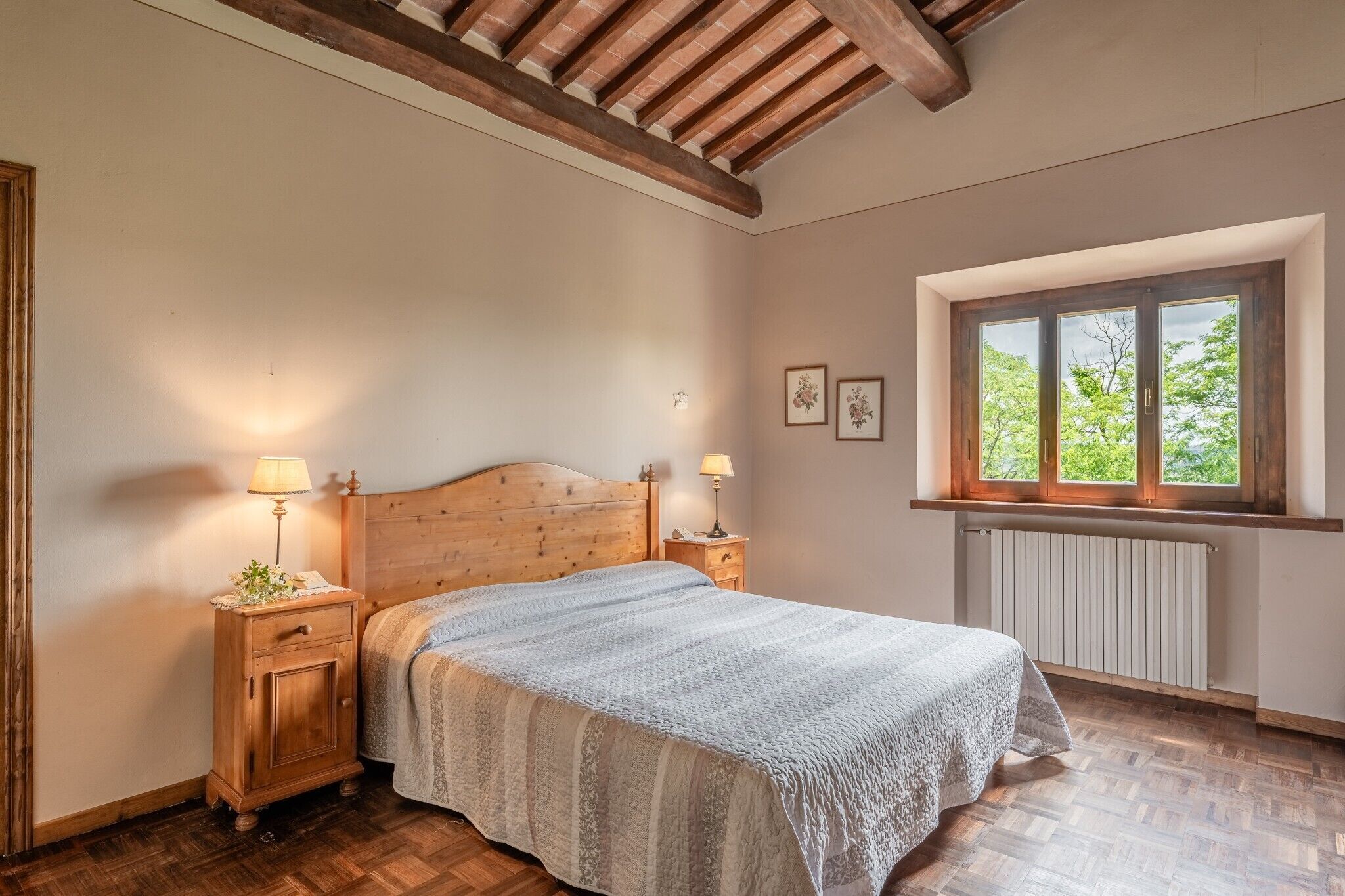Une chambre à coucher avec une tête de lit en bois, un lit recouvert d’une couvre-lit à motifs, des lampes de chevet, une fenêtre donnant sur des arbres et un radiateur.