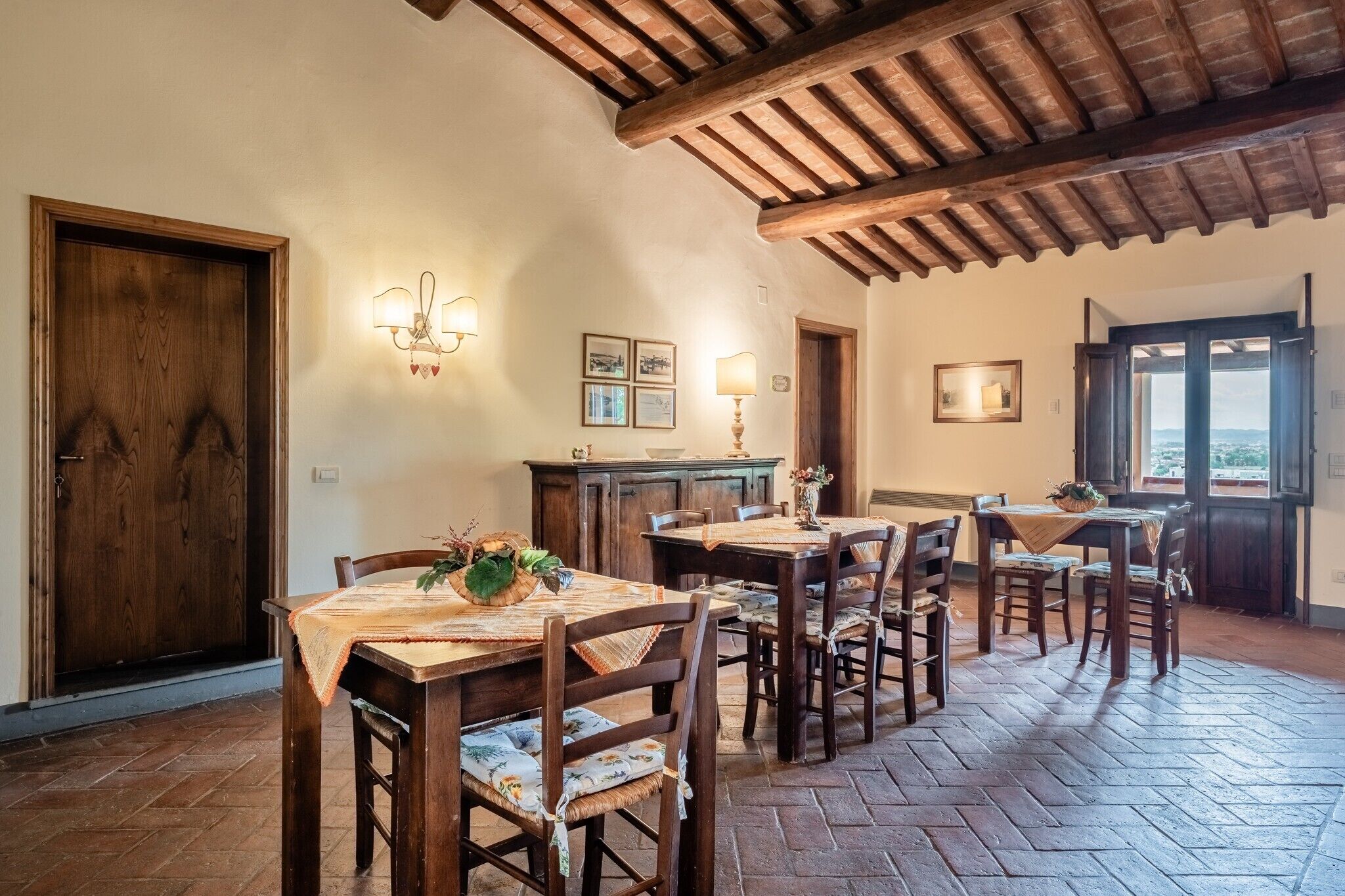 A dining area with wooden tables and chairs, a wooden ceiling, and a view through an open door.