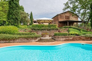 Une piscine bordée de briques, un mur en pierre et un bâtiment à la façade en briques surmontée d’un auvent blanc.