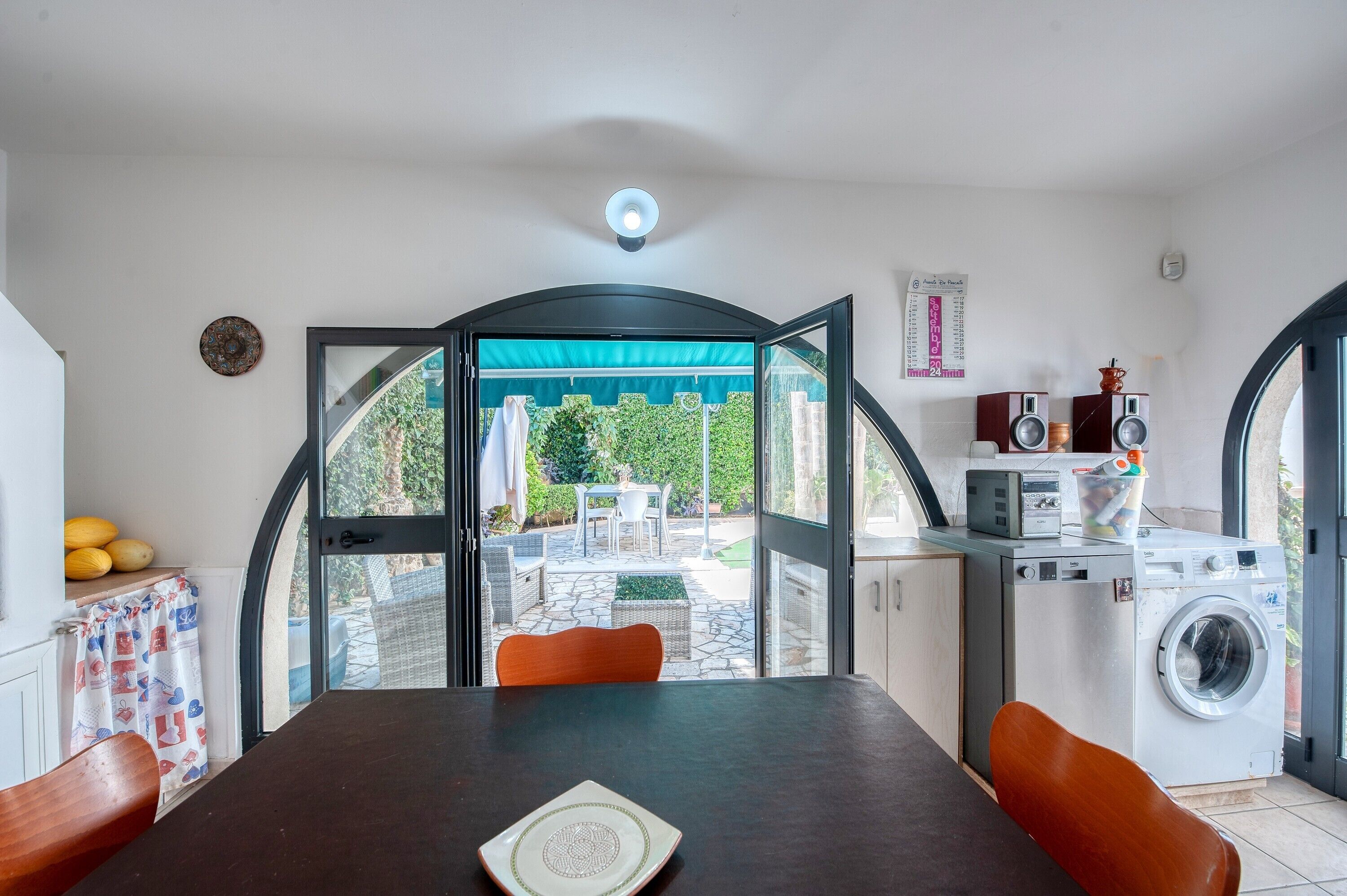 A room with a table, chairs, a washing machine, speakers, and a view of a garden.