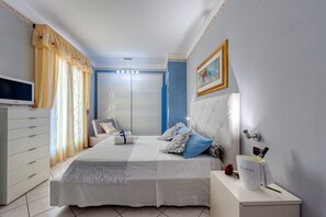 Un dormitorio con cama, mesitas de noche, televisión, cortinas, un cuadro y un armario.