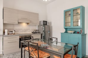 A compact kitchen with a glass dining table, chairs, a teal cabinet, and a microwave on the counter.